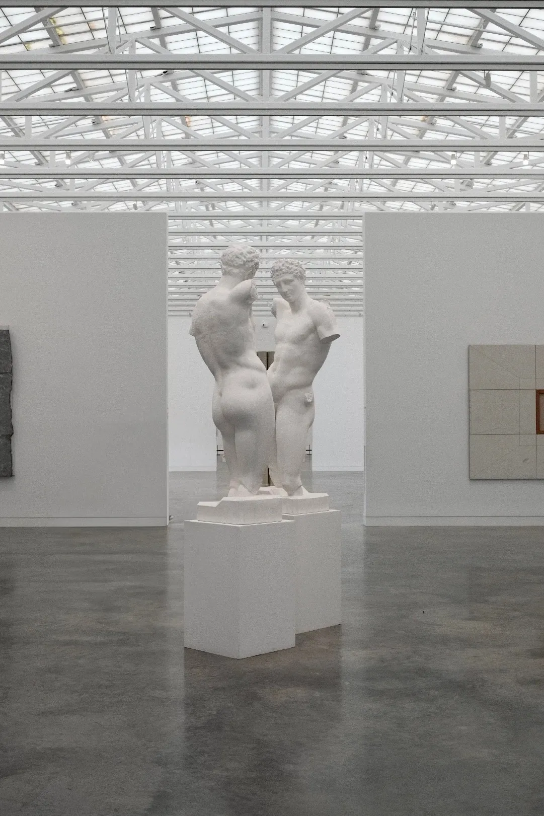 Two white plaster figures on plinths in a skylit gallery with a concrete floor